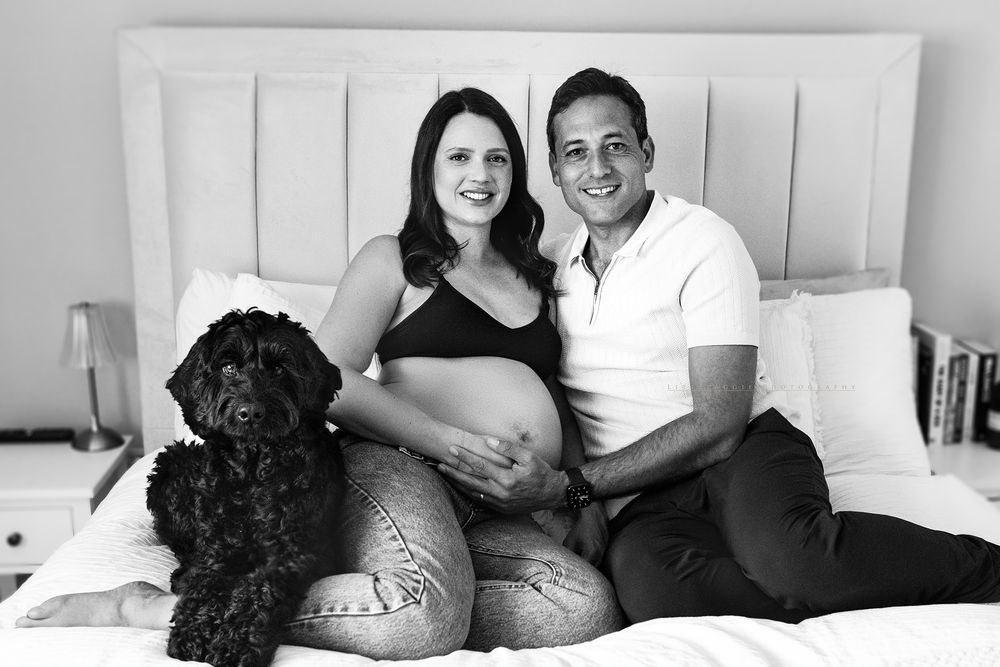 Black and white maternity photo of a couple sitting on a bed with their black doodle dog, woman cradling baby bump.