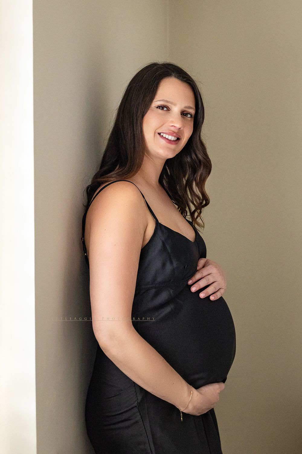 Smiling pregnant woman in black satin dress cradling baby bump against neutral wall in maternity photo.
