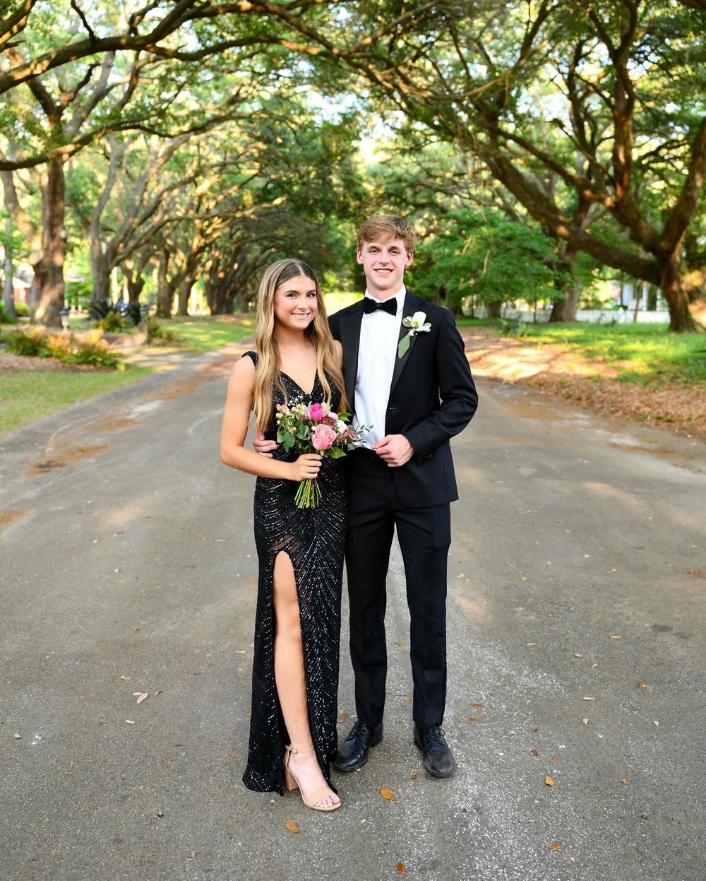 Formal couple poses on oak-lined path, woman in black gown with pink bouquet.
