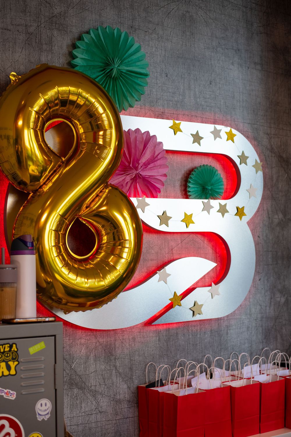 Gold number 8 balloon beside illuminated letter B with stars, colorful paper fans, and red gift bags at a birthday party.