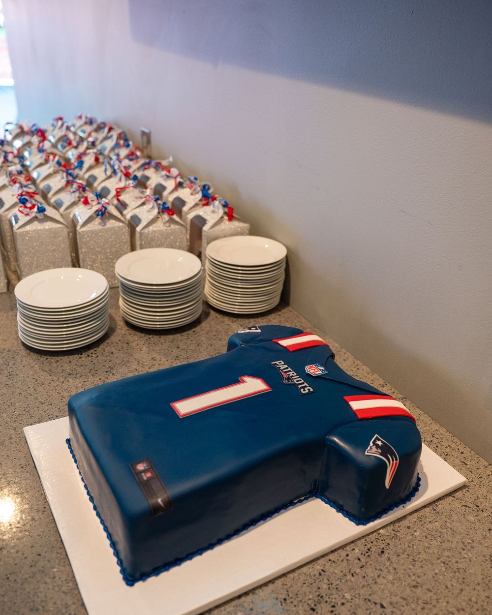 New England Patriots jersey-shaped cake in blue fondant with number 1, displayed on a table with stacked plates and decorated cupcakes.