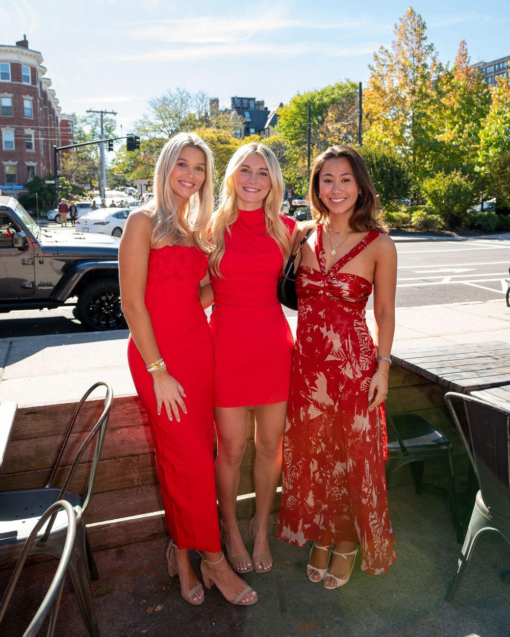Three women in red dresses smiling together on a sunny outdoor patio in an urban setting.