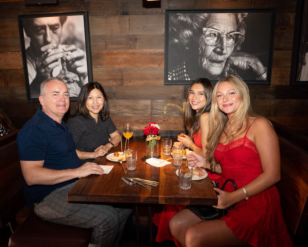 Four people smiling and dining together at a restaurant table with cocktails and artwork on dark wood walls.