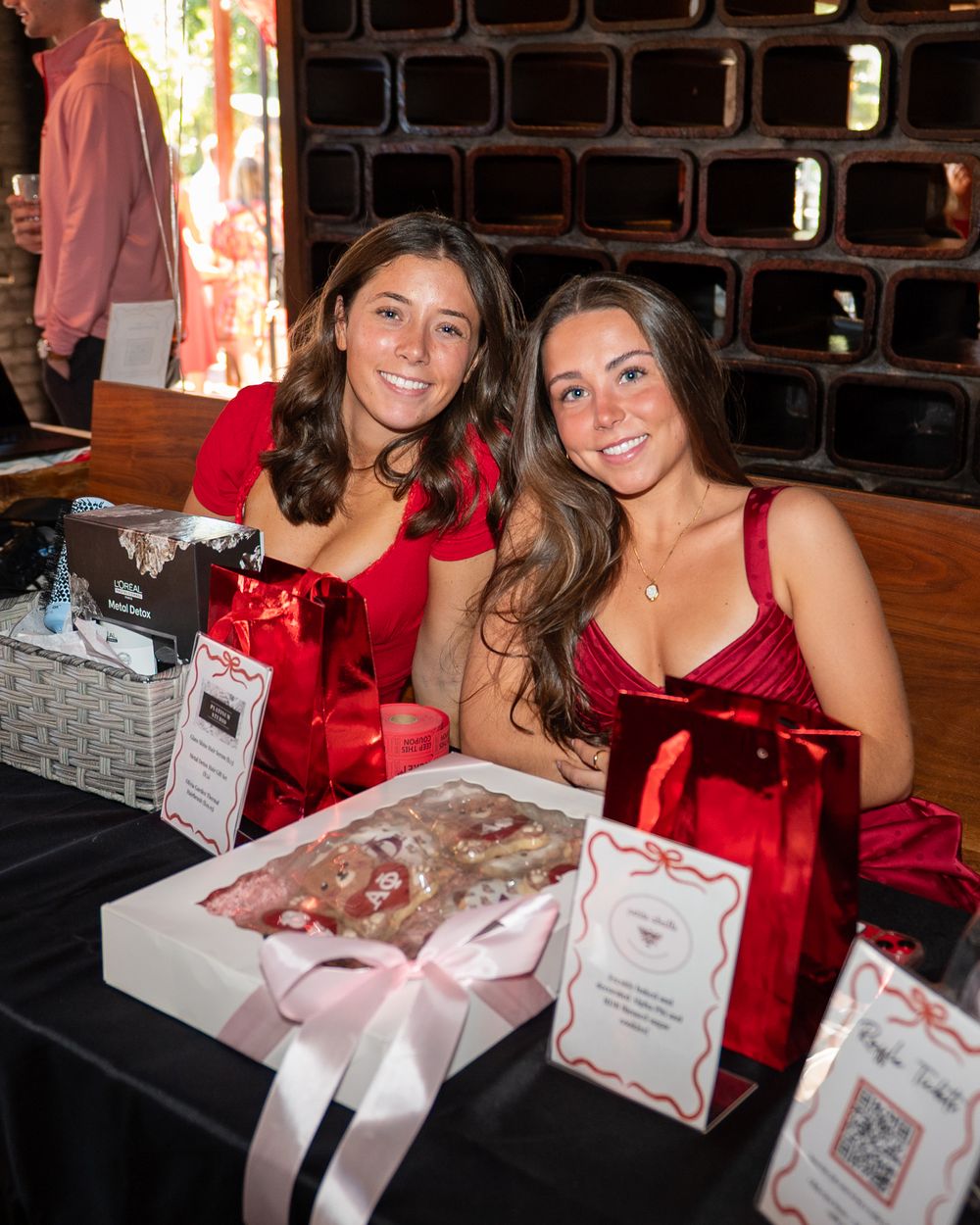 Two smiling women in red dresses sit at a decorated table with gift boxes and bags at a festive event.