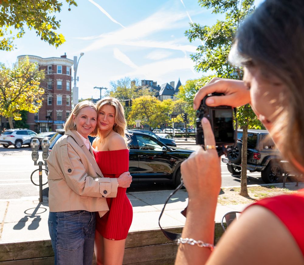 Two women pose for a photo outdoors while a third woman photographs them with a camera on a sunny day.