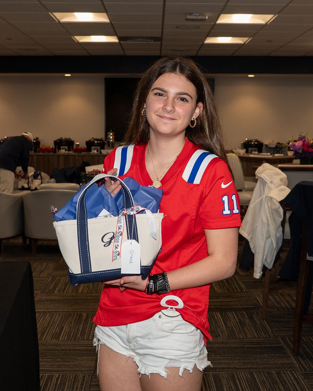 Young woman in a red Patriots jersey holding a navy and white tote bag at an indoor event.