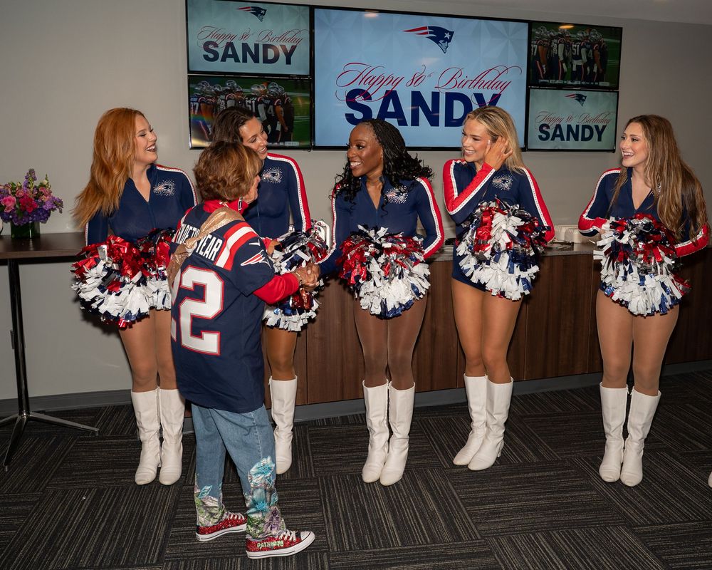 New England Patriots cheerleaders celebrate Sandy's birthday in a suite, posing with a fan wearing a Patriots jersey.