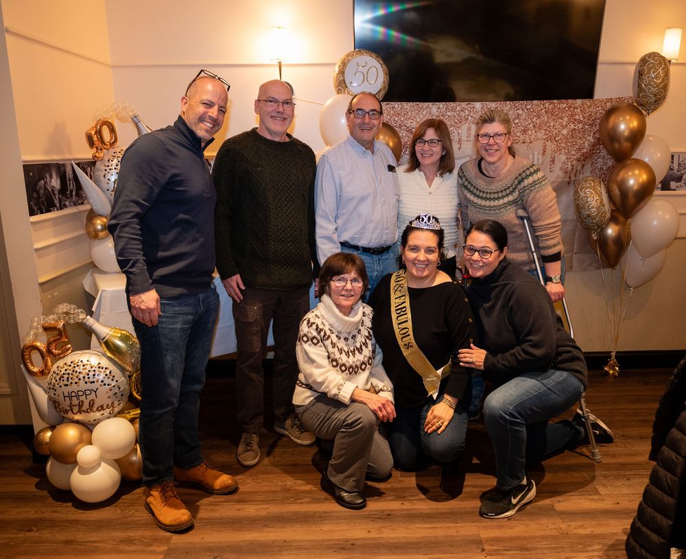 Group of eight people posing together at a milestone birthday party with gold and white balloon decorations.