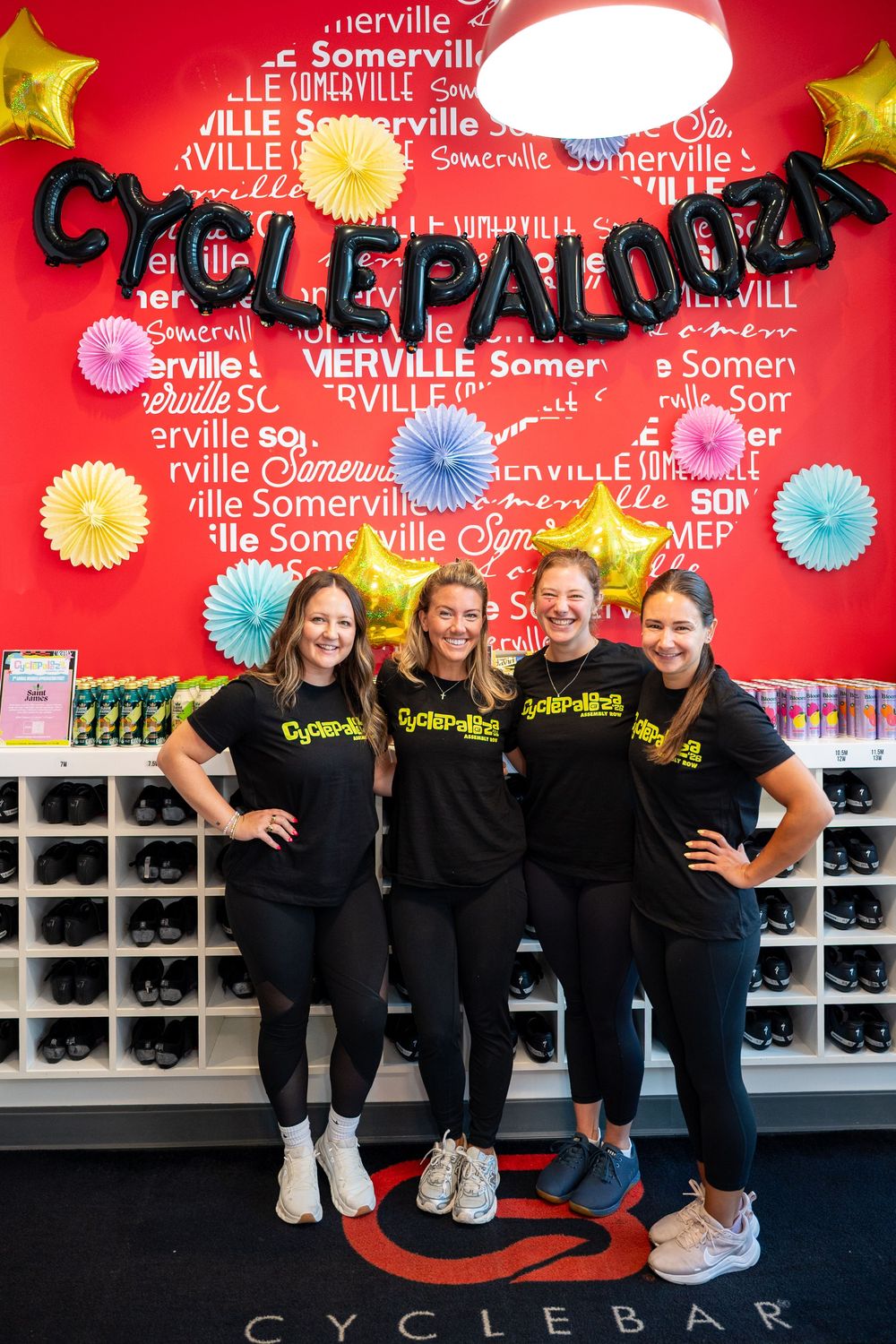 Four women in Cyclepalooza shirts pose at CycleBar Somerville with colorful decorations and balloon letters.