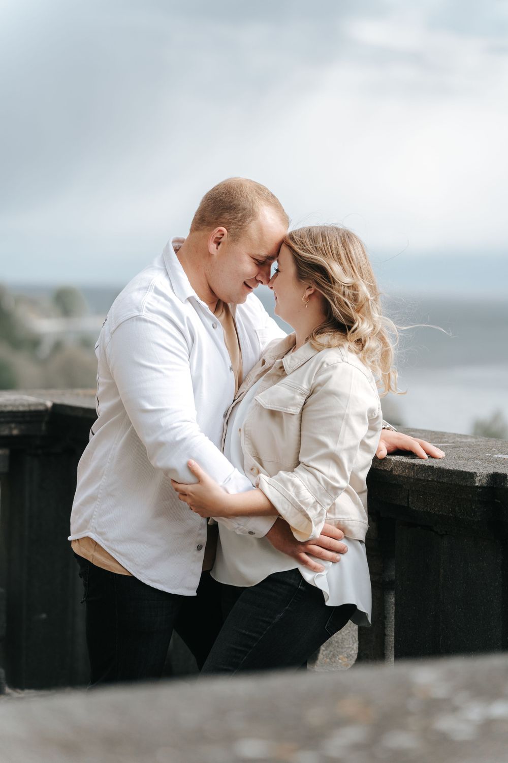 A romantic moment between a couple in white clothing leaning against a dark railing by the sea.