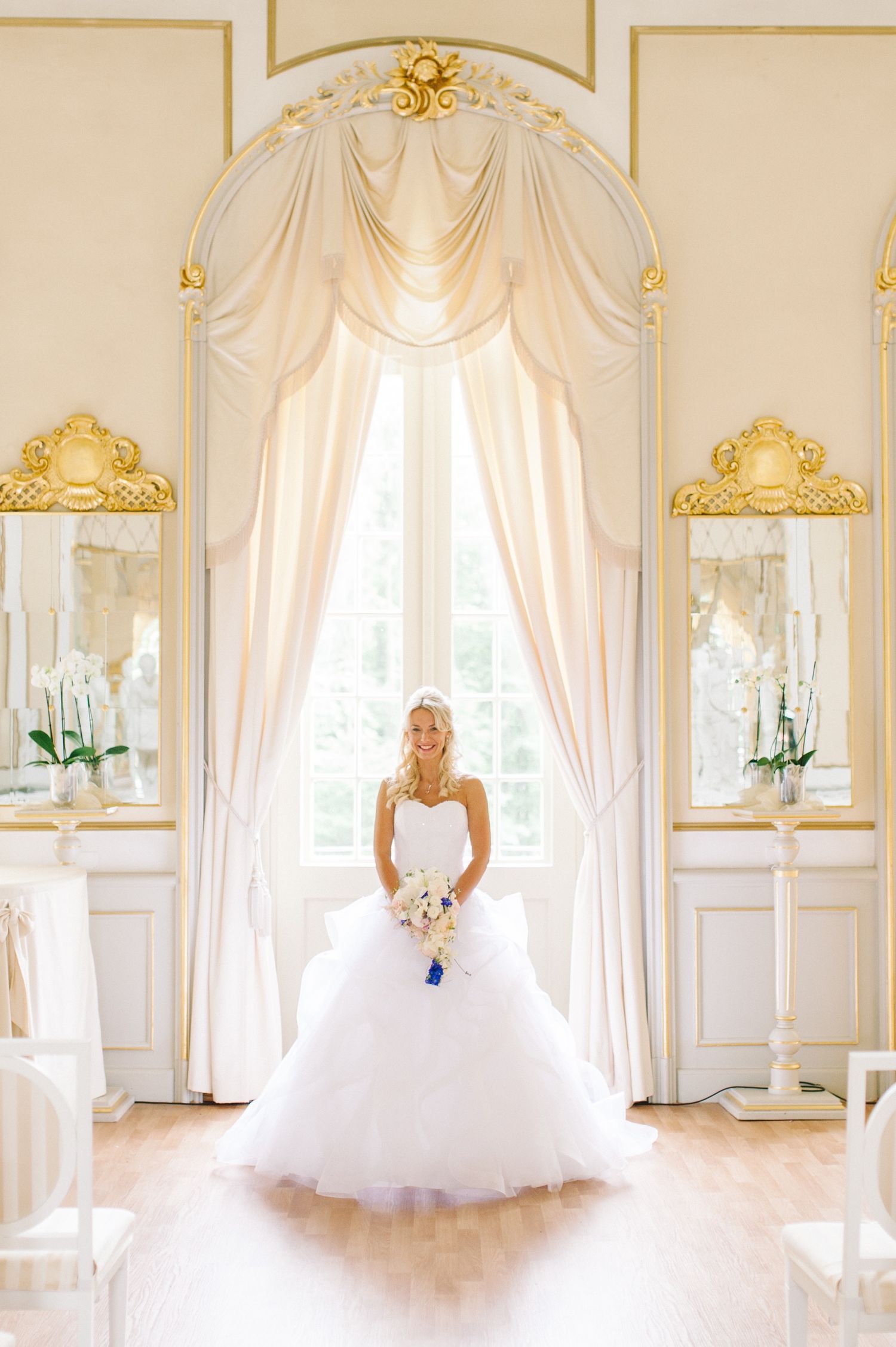 A bride in a white gown poses dramatically at an elegant window with ornate gold mirrors. Organized wedding by MyCastleWedding | Dresden.