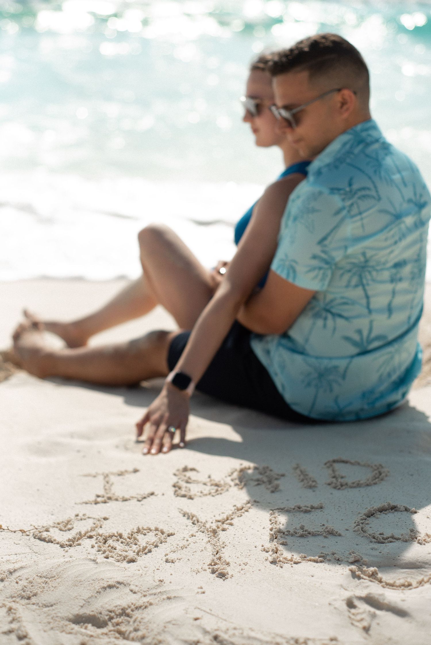 Jola and Bruno sitting on the beach of Cancun taking their engagement photos, on the sand is written "I said yes"