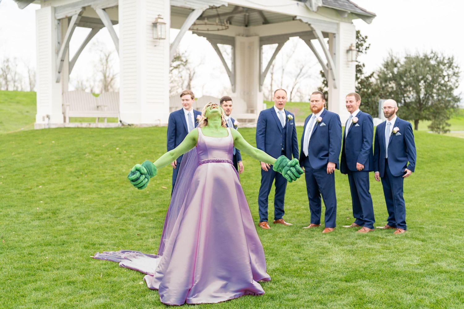 Wedding party poses under a white pergola with a person in purple gown standing in front.