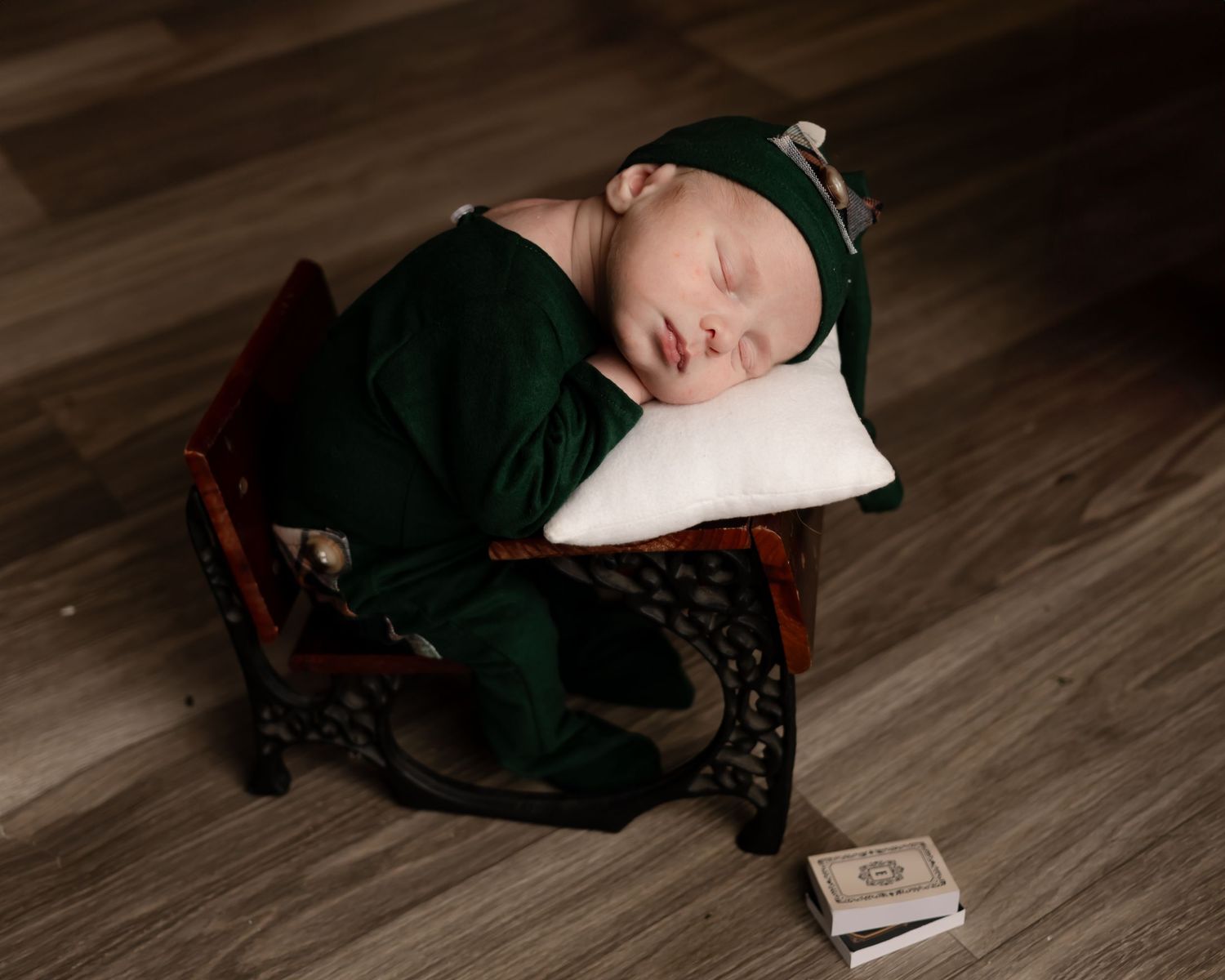 Newborn in dark green outfit sleeping peacefully in wooden chair with small book nearby.