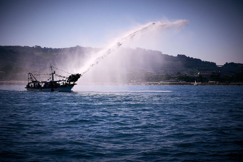 Silhouetted fishing boat sprays water from its side in front of coastal hillside.