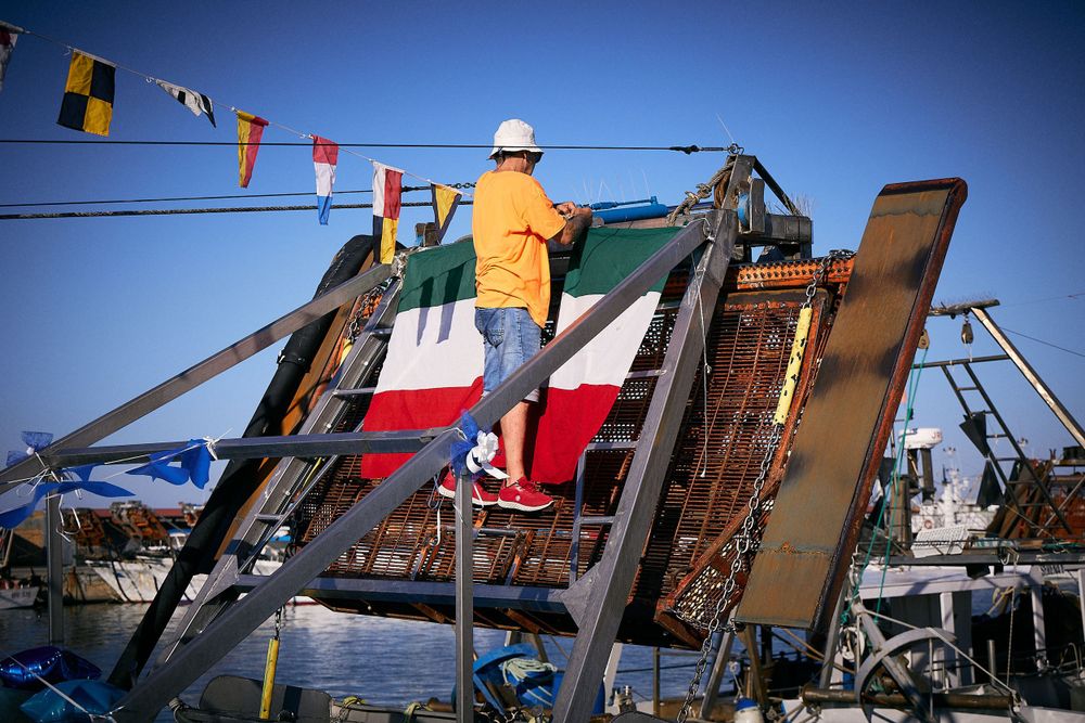 Worker in orange safety vest performs maintenance on equipment with flags visible against blue sky background.