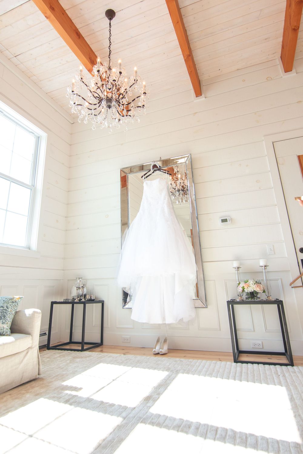 A bright white interior room with exposed wooden beams, large windows, and elegant chandeliers casting natural light with a wedding gown