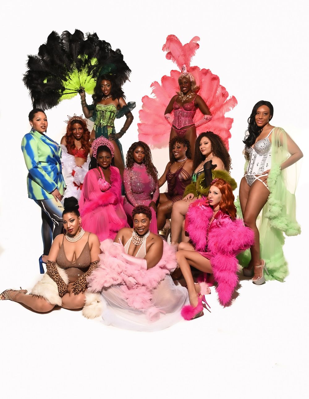 A glamorous group poses together wearing colorful feathered costumes and outfits in pink, green, and black against a white backdrop.