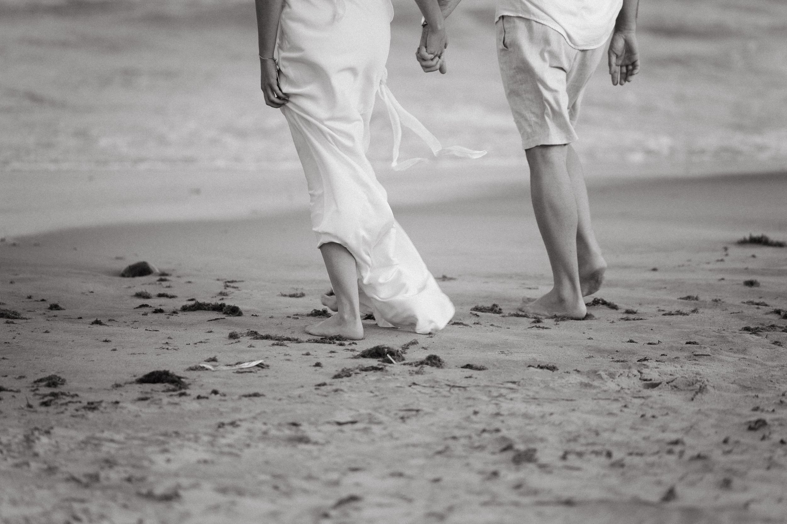 Couple walking barefoot on sandy beach holding hands in black and white.