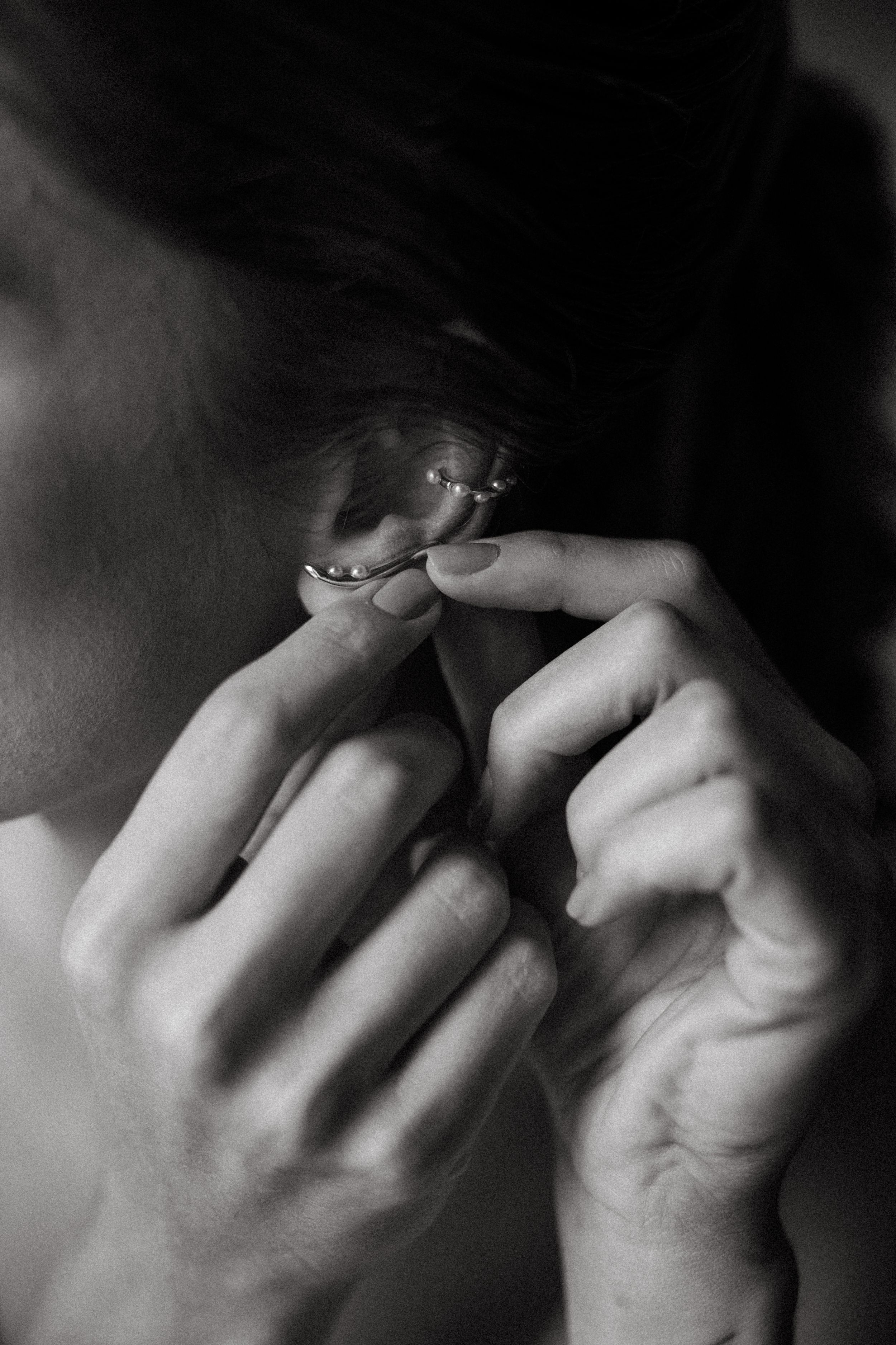 Black and white close-up of a woman putting on a delicate ear cuff jewelry piece with both hands.