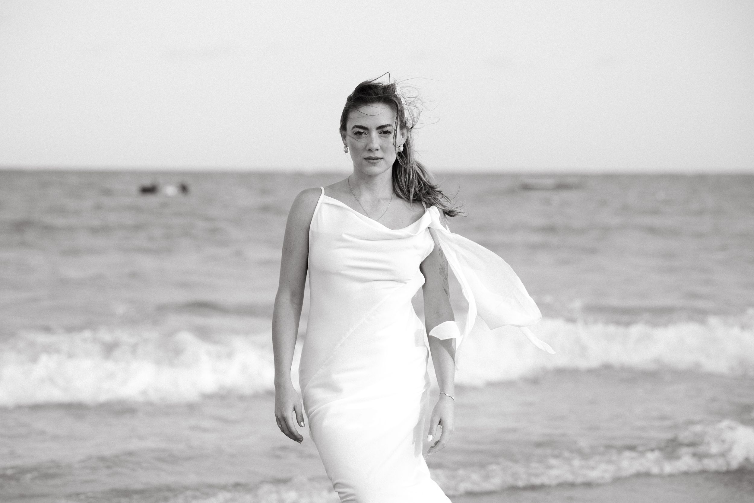 Black and white portrait of elegant woman in white slip dress walking along ocean shoreline with waves.