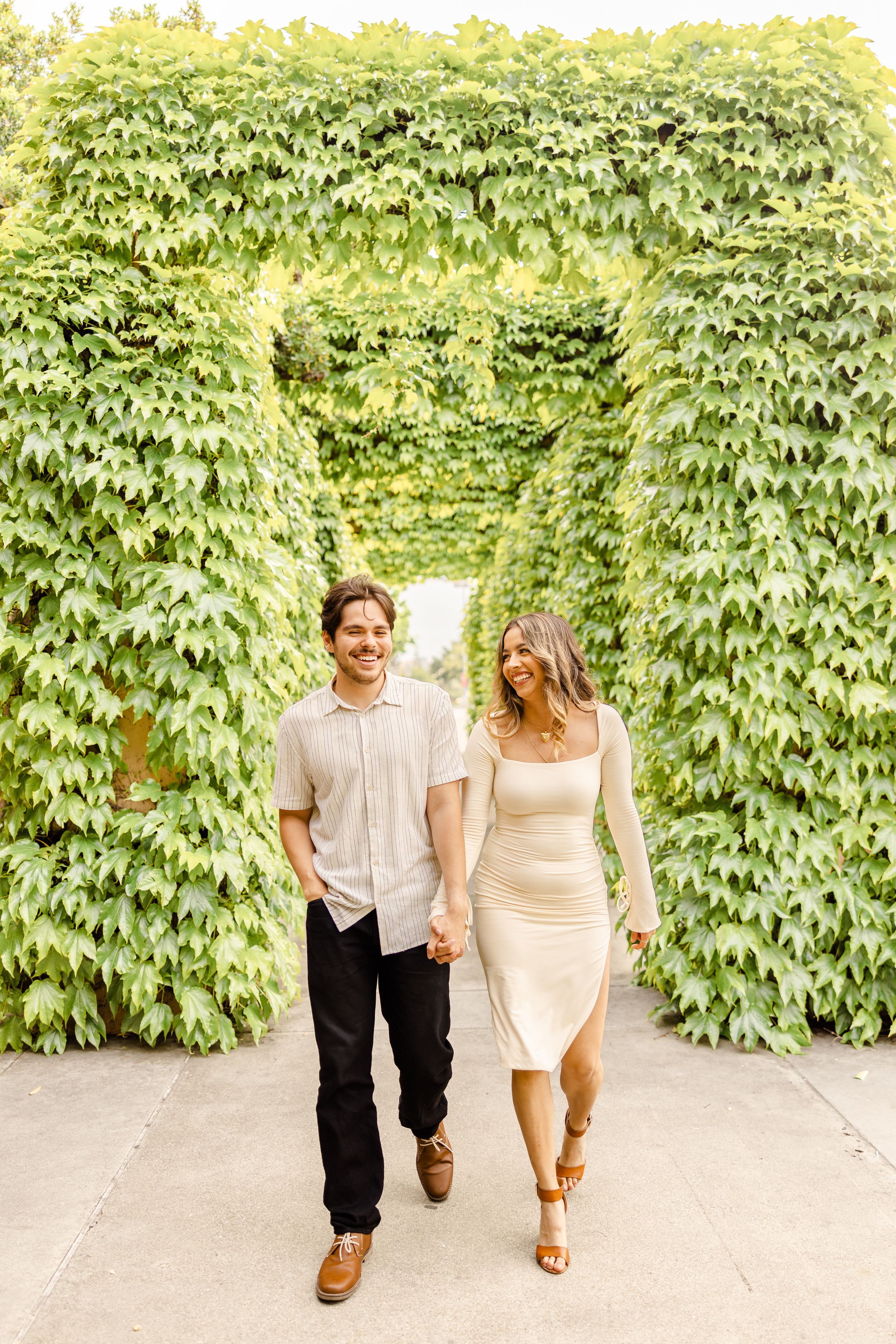Couple walks hand in hand along ivy-covered pathway in casual attire.