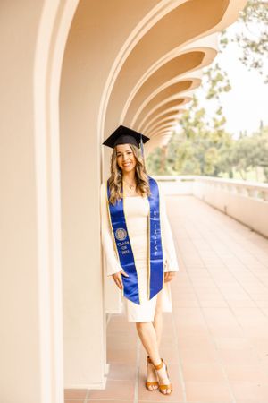 Graduate in white dress and blue stole poses under arched walkway during graduation photoshoot.