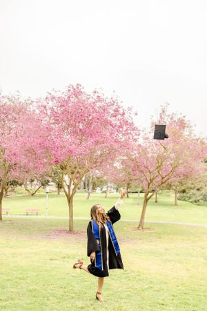 Graduate celebrates under blooming cherry blossom trees by tossing cap in the air.