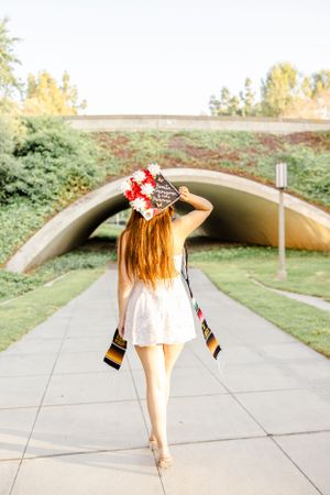 Graduate in decorated cap and stole twirls on pathway near concrete tunnel structure.