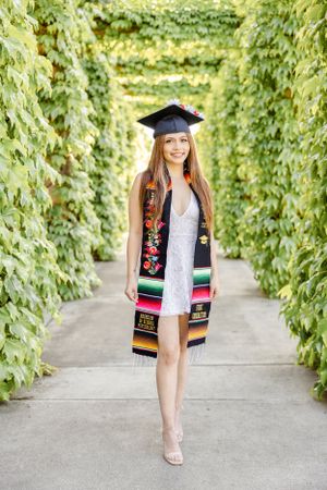 Graduate in cap and colorful stole poses in lush green vine-covered walkway.