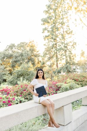 Graduate in white dress poses on concrete steps with blooming flowers in background.