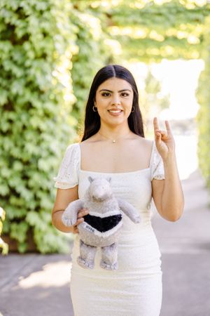 Graduate in white dress holds stuffed animal while posing outdoors.