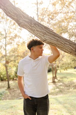 Student in white shirt leans against tree trunk in natural outdoor setting.