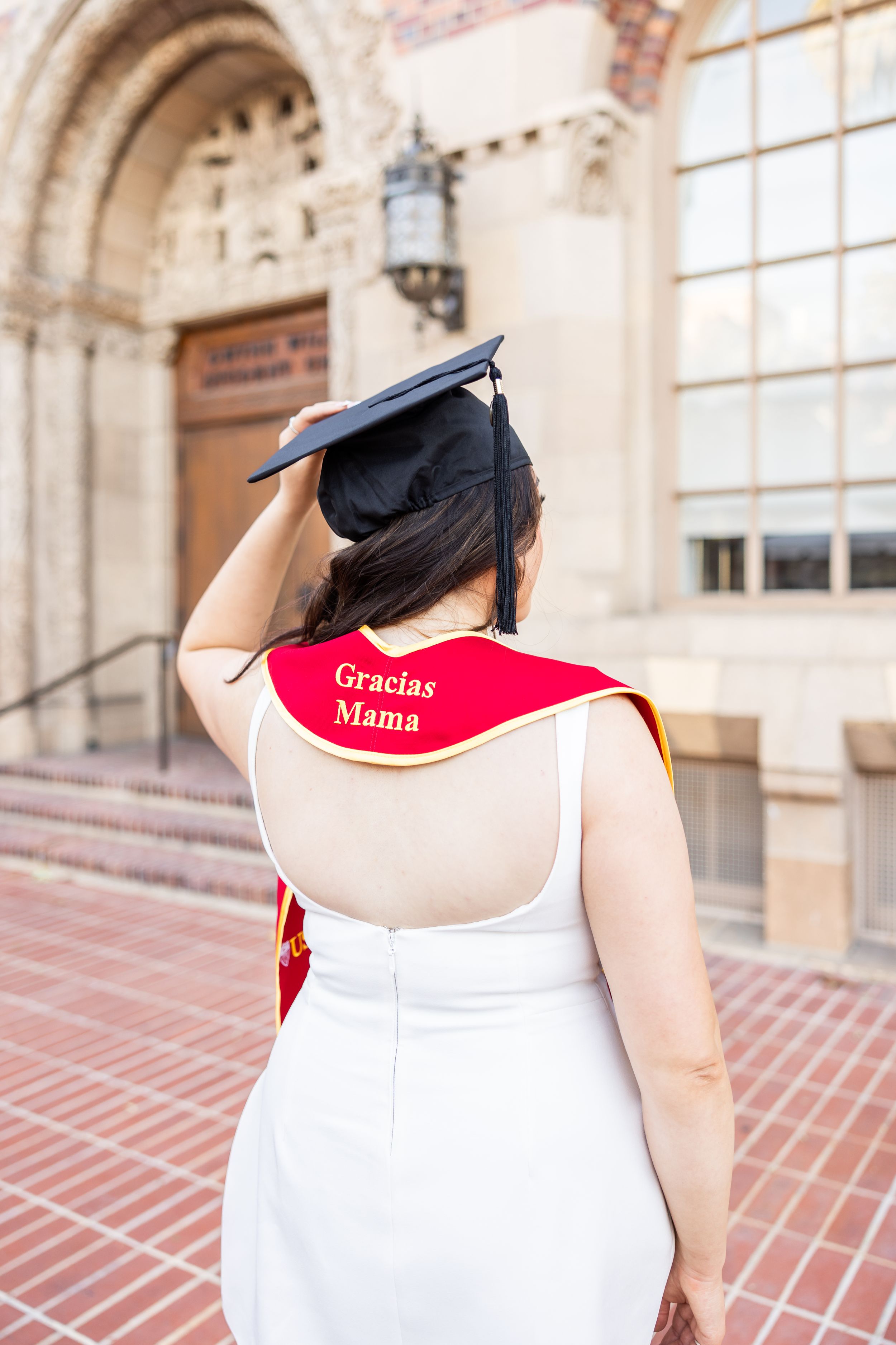Graduate wearing cap and white dress with personalized red stole on university steps.