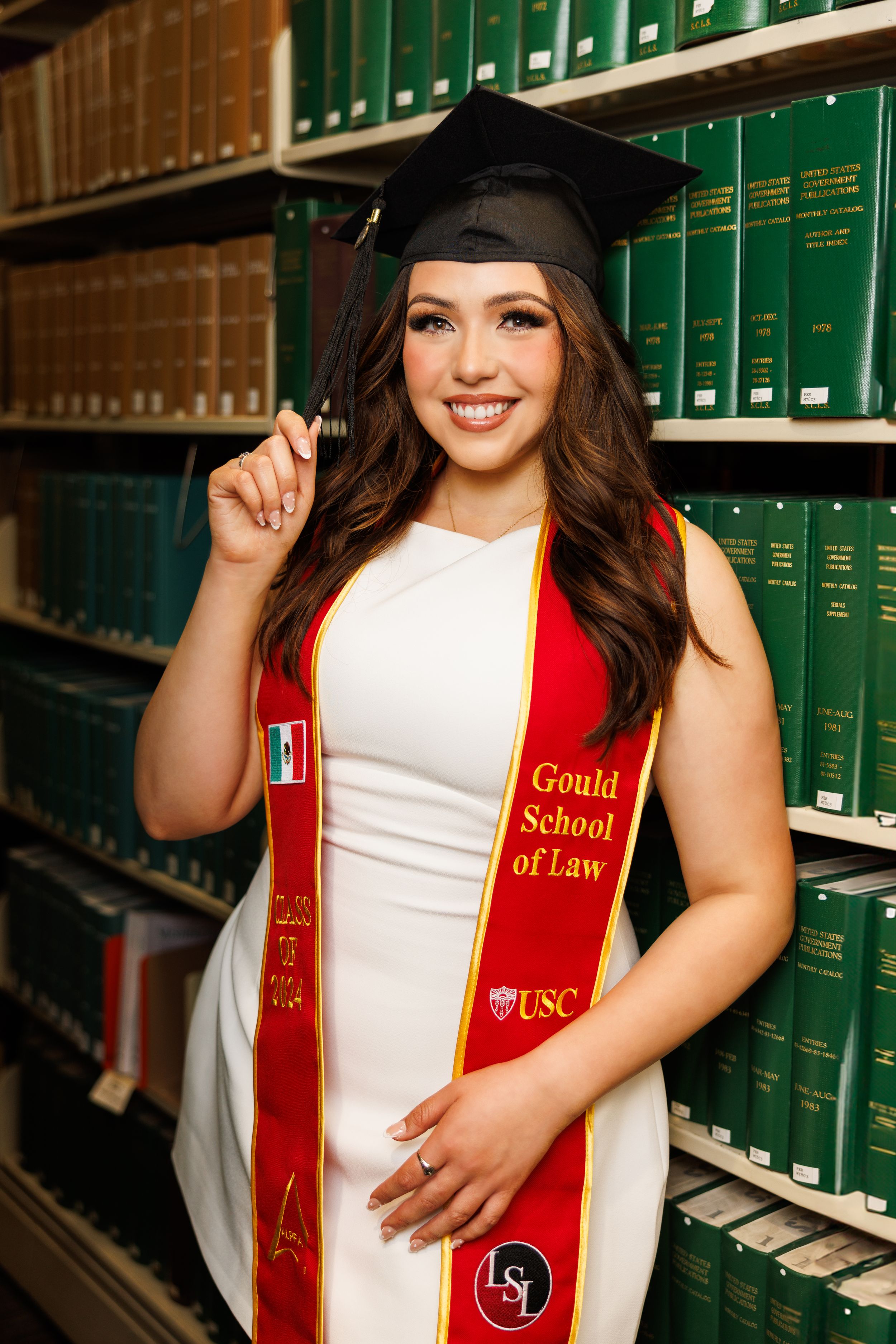 Graduate in white dress and red stole poses in law library with green books in background.