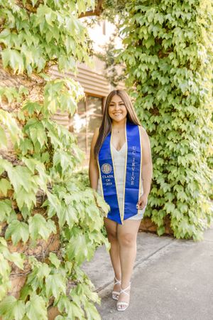 Graduate in blue sash and cap poses in front of lush green ivy backdrop.