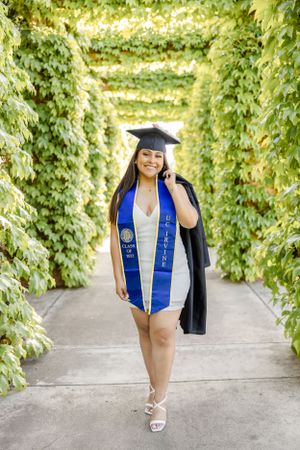 Graduate in academic regalia poses in front of ivy-covered wall outdoors.