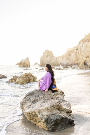 Woman relaxing on large coastal rock with purple blanket by the ocean.