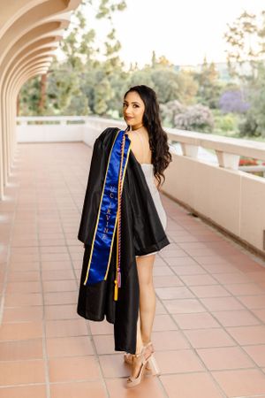 Graduate in black cap and gown with blue sash poses in an outdoor corridor with terra cotta tiles.