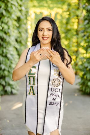 Graduate in white dress displays UCI Class of 2023 sash in garden setting.