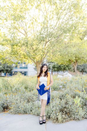 Graduate in yellow cardigan and blue sash poses outdoors among blooming spring foliage.