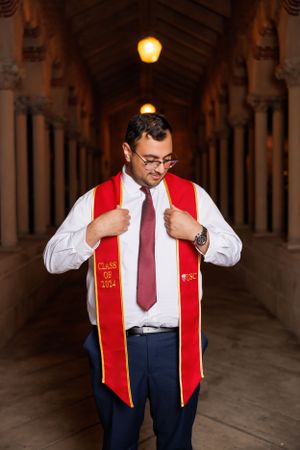Graduate adjusts red graduation stole while standing in grand architectural corridor.