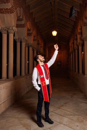 Graduate in red stole points upward while standing in historic columned hallway with dramatic lighting.