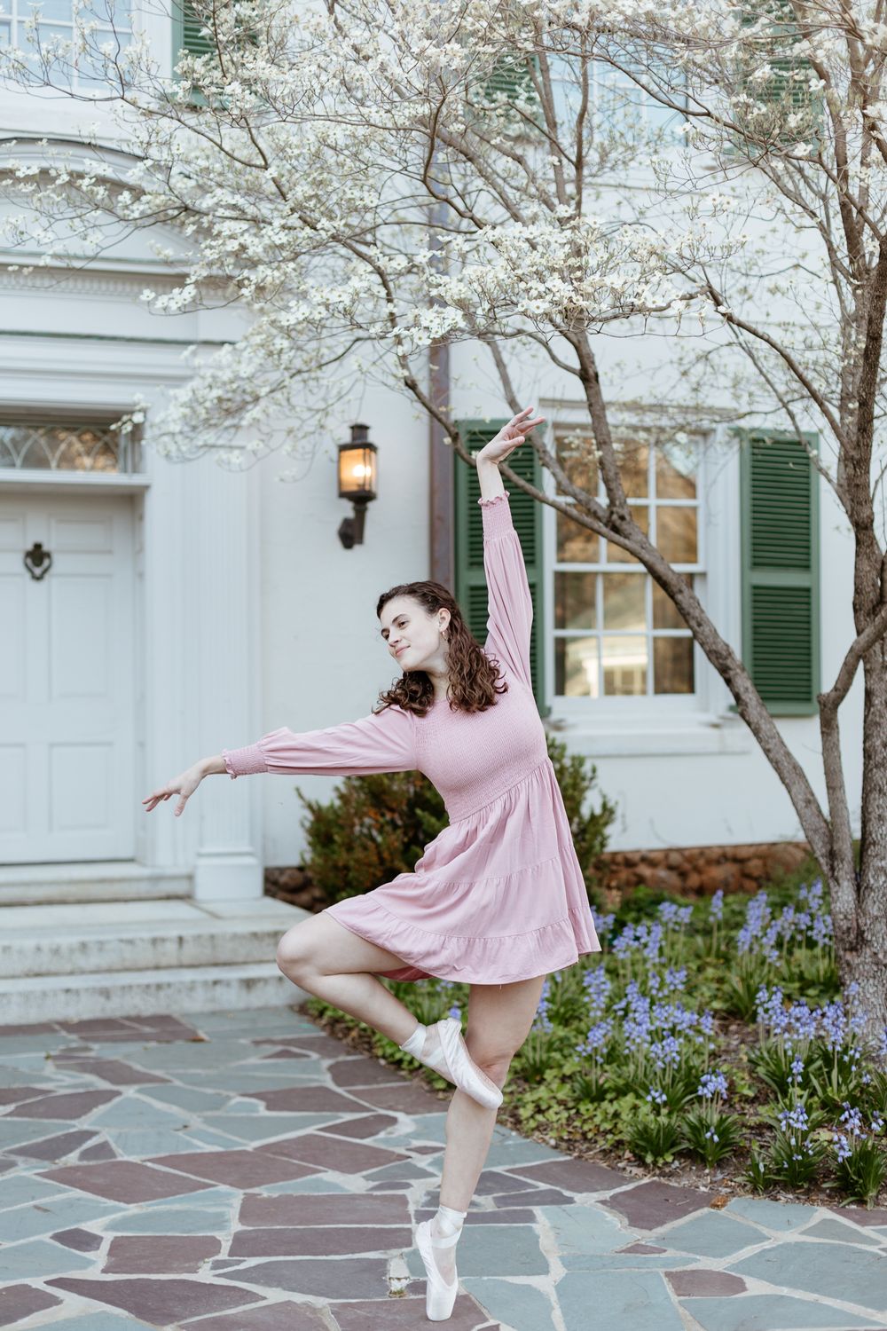 Ballet dancer performs elegant pose in front of white building with flowering trees.