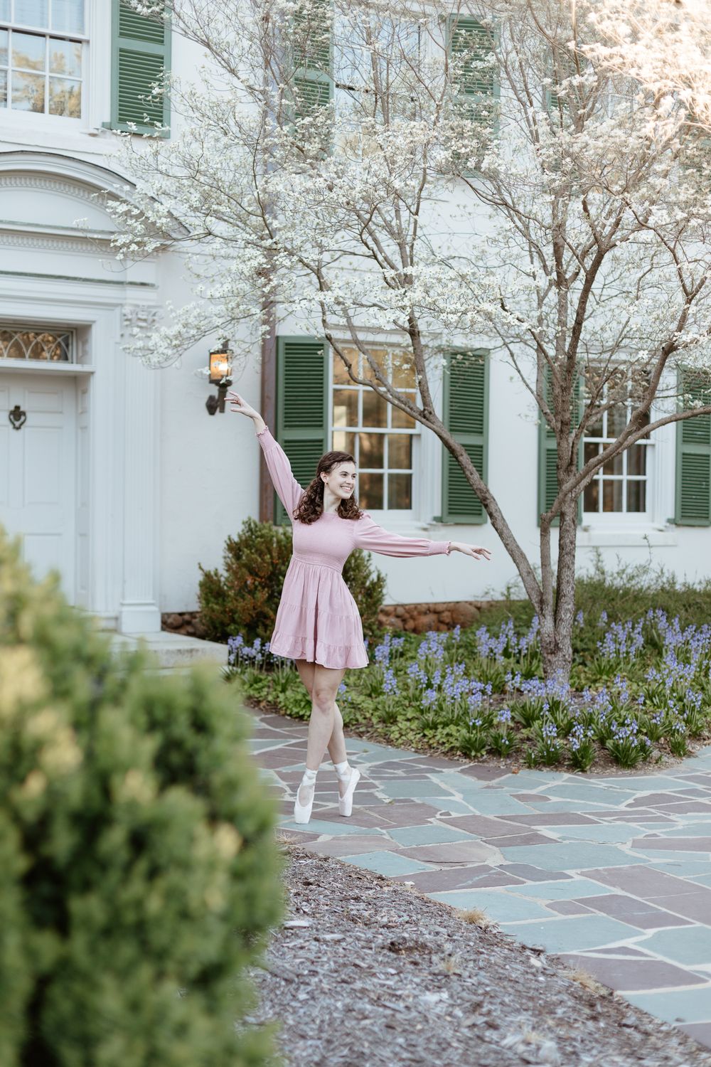 Ballerina strikes graceful pose on stone patio with blooming trees in background.