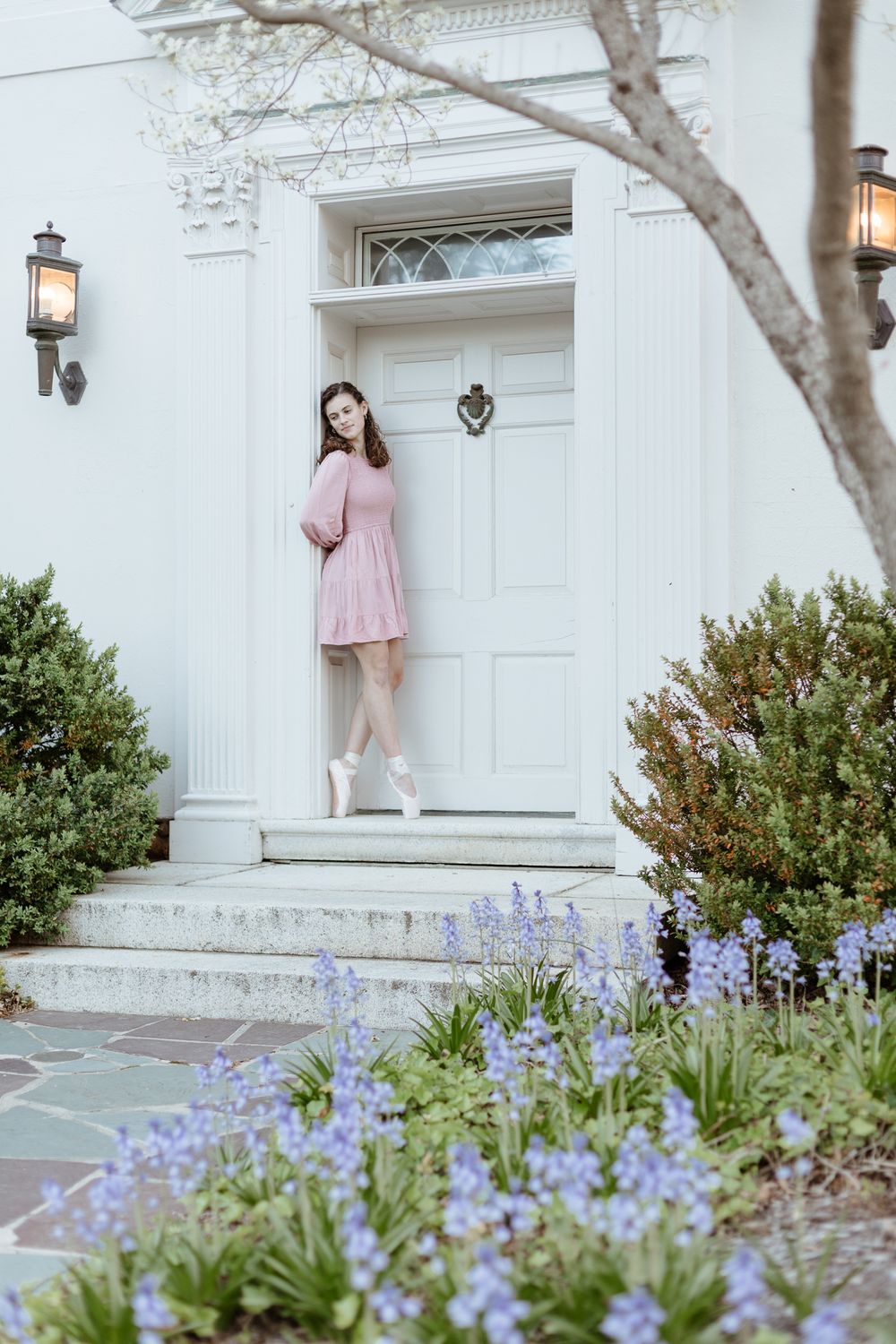 Pink-dressed dancer poses by white door with purple flowers blooming in garden.