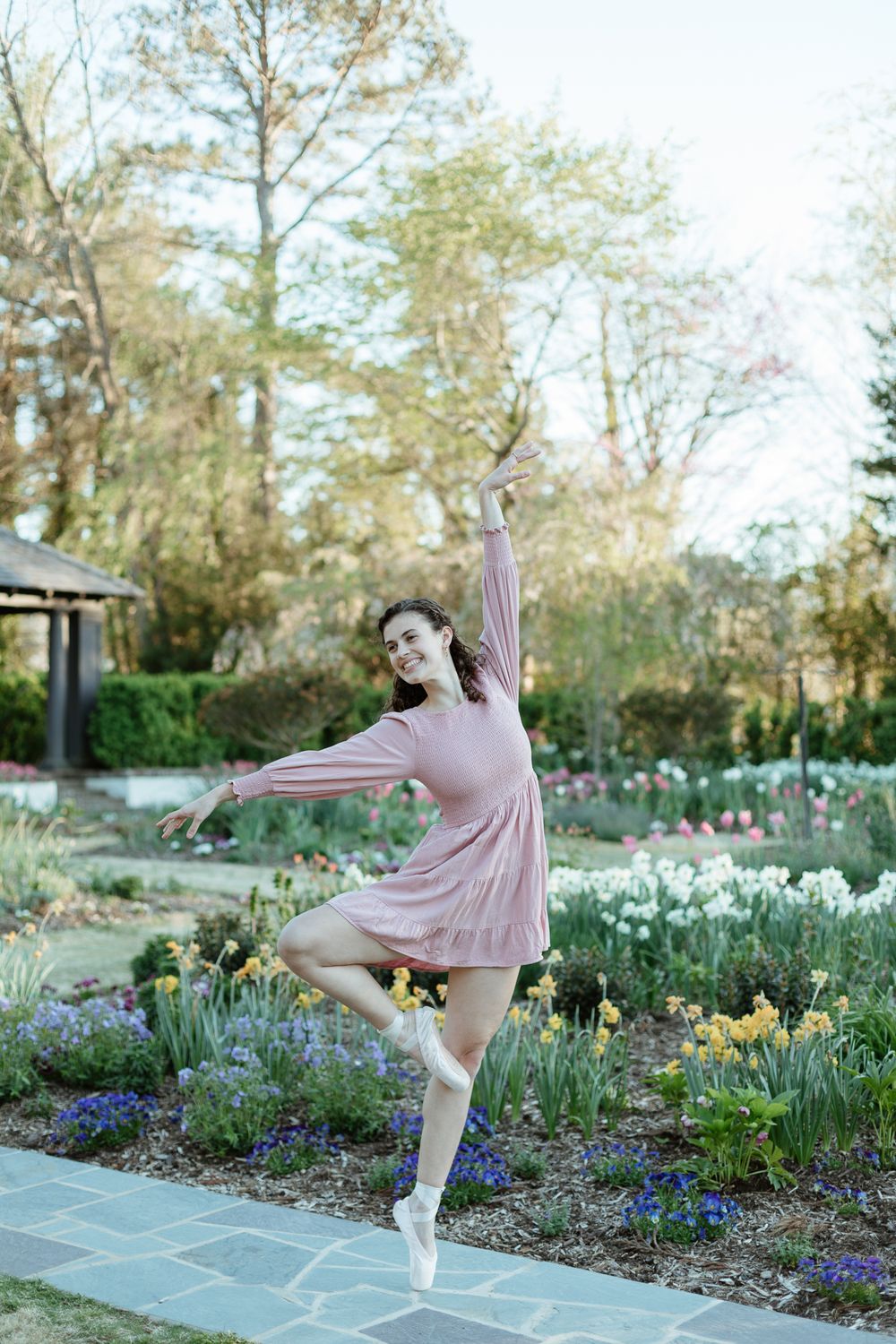 Ballet dancer performs graceful movements in garden filled with spring daffodils and purple flowers.
