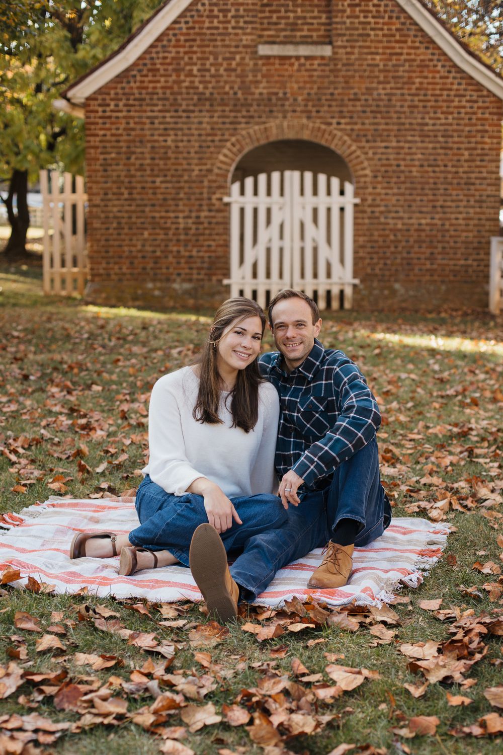 A couple relaxes on a picnic blanket surrounded by fallen autumn leaves near a brick building.