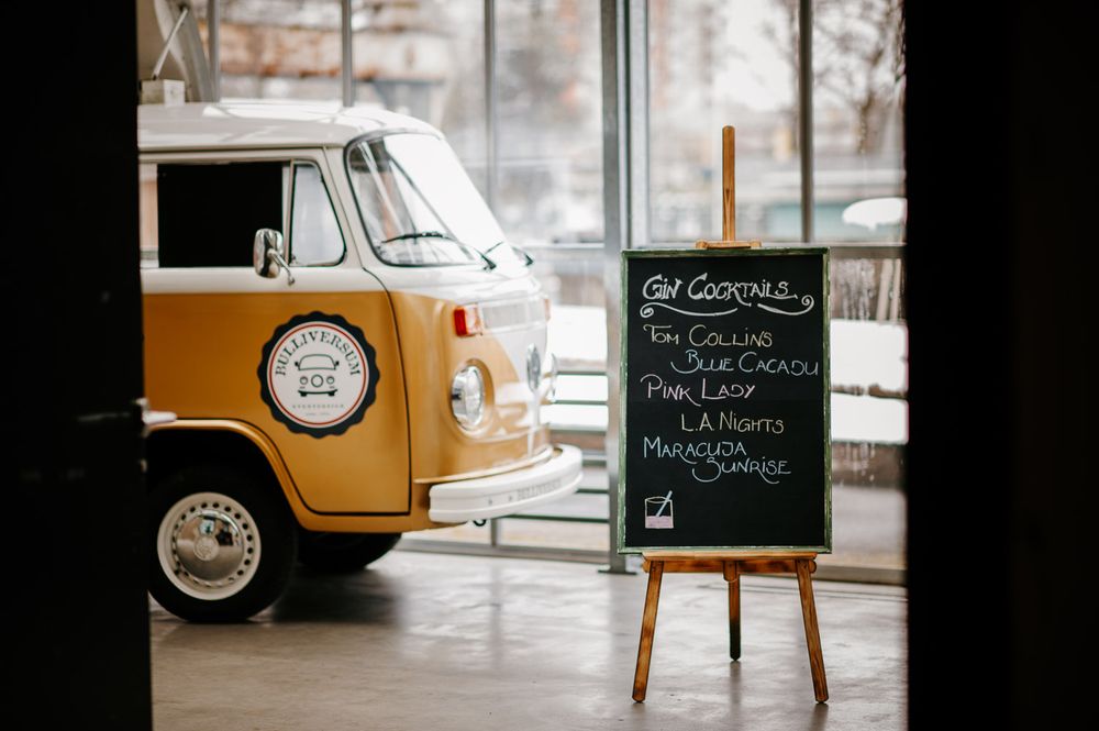 A yellow VW camper van beside a chalkboard cocktail menu sign at an indoor event.