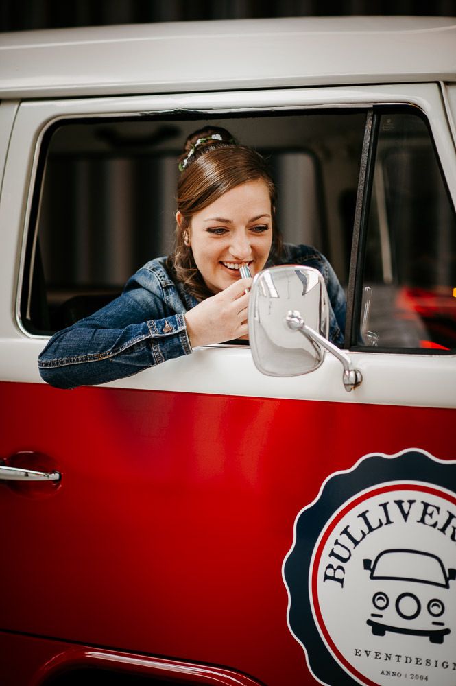 A smiling bride in a denim jacket looking into the mirror of a red and white VW camper van.